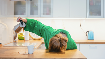Müde Frau mit dem Kopf auf dem Tisch, die den Kaffee neben die Tasse schüttet.